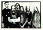 Mustavalkoinen valokuva At The Gates -bändin kuudesta miesjäsenestä, jotka seisovat valkoista lautaseinää vasten. Osalla pitkät hiukset, osalla partaa ja viikset.