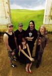 Viisihenkisen Shattered Within -bändin jäsenet seisovat hietikolla vehreän nurmikon edustalla. Kuvakulma hieman ylhäältä alaspäin. Kuvassa neljä miestä, jotka seisovat naisen takana.