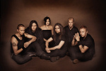 Tummanruskea valokuva Hevein-bändin jäsenistä, jotka istuvat rykelmänä maassa. Heitä on viisi miestä ja yksi nainen. Nainen istuu miesten takana keskimmäisenä. Osalla pitkät hiukset, osalla lyhyet.