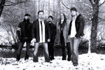 Sinisävyinen ja kylmänoloinen valokuva Winterborn-bändin miehistä, jotka seisovat ulkoilmassa lumisen maan ympäröimänä. Taustalla lehdettömiä puita ja joki. Kuvassa viisi miestä, joista keskimmäisellä yllään musta takki, kuluneet farkut ja valkoinen kaula