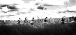 Mustavalkoinen ja hieman ylivalottunut valokuva Mokoma-bändin miehistöstä, joka seikkailee keskellä korkeana kasvavaa ruohikkoa tai peltoa pilvisen ja kirkkaana hohtavan taivaan alla. Miehillä on käsissään soittimia, kitaraa, bassoa.