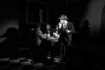 Mustavalkoinen promovalokuva Twiggy Oliverista, joka istuu pienen pöydän ääressä hattu päässään. Pöydän ympärillä istuu kaksi muuta miestä. Huone on hyvin hämärä.