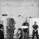Harmaavalkosävyinen valokuva Dark Filth Franity -bändin jäsenistä, jotka seisovat ulkoilmassa suuren tiilikiviseinän edustalla. Miesten seassa seisoo kaulan korkeudella katki sahattu tammi tai muu puu. Miehistä fototssa neljä kapapletta. Osalla mustat vaa