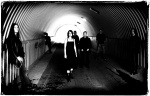 Harmaasävyinen valokuva Silentium-bändin kokoonpanosta, joka seisoo ulkoilmassa pitkän puolikaarimaisen tunnelin suuaukolla. Tunnelin toisessa päässä valoa. Kuvassa lauma mustiin pukeutuneita pitkähiuksisia miehiä ja heidän keskellä mustaan mekkoon pukeut