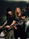 Children Of Bodom -yhtyeen Aleksi Laiho ja kitaristi irvistelevät mustahkoa taustaa vasten. Miehet seisovat instrumentit kourassa vieretysten. Kuvan oikeassa laidassa ja yläosassa näkyy vaaleampaa valoa tai savua.