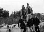 Harmaasävyinen ryhmäkuva Adastra-bändin kokoonpanosta. Miehistö seisoo ulkosalla suurten betonikierukoiden päällä. Kuvassa viisi miestä, joista kolmella alarivin miehellä mustat vaatteet ja kahdella taaemmalla harmaat takit ja vaaleampaa vaatetta yllään.