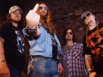 Doom metal -bändi Orange Goblinin tiimi seisoo sikin sokin yllään arkivaatteet. Keskimmäisellä miehellä aurinkolasit silmien edessä ja yllään sininen farkkutakki ja farkkuhousut. Hän näyttää kohtisuoraan keskisormea. Kuvassa yhteensä neljä miestä.
