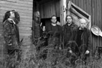 Harmaasävyinen valokuva Spiritus Mortis -bändin kokoonpanosta. Jäsenet seisovat puuseinän edustalla ruohikossa. Heillä mustaa vaatetta yllään. Osalla pitkät hiukset, osalla kalju, kaikki ovat miehiä.