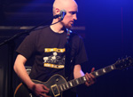 Valokuvassa FM2000:n kaljupäinen mies soittamassa kitaraa tai bassoa tummansinisen valon hohtaessa taustala. Mies katsoo vasemmalta oikealle ja pitää instrumenttia kummallakin kädellä kiinni.