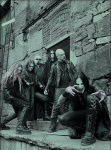 Dimmu Borgir -bändin jäsenet savenharmaassa kuvassa. Shagrath irvistelee ja näyttää käsimerkkejä. Muut bändin jäsenet seisovat hieman taaempana. Kuvan oikeassa laidassa kivinen seinä, jossa suuaukko.