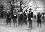 Harmaasävyinen valokuva mustiin pukeutuneista ihmisistä, joista osa miehiä ja osa naisia. Kuvassa yhteensä kuusi heppua, jotka seisovat rivissä ulkoilmassa lehdettömien puiden edustalla. Kuvan alaosassa valkoista utua.