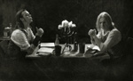 Tummasävyinen ja harmaasävyinen valokuva kahdesta miehestä, jtoka istuvat mustaa taustaa vasten pöydän ääressä toisiaan vastapäätä. Pöydällä on keskellä kynttelikkö ja papereita.