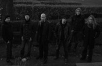 Lauma miehiä seisoo mustiin pukeutuneen yhdessä rivissä pensaiden edustalla tummassa ja harmaasävyisessä valokuvassa.