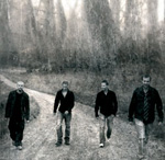 Lähes harmaasävyinen valokuva tiellä rivissä kävelevistä neljästä miehestä. Heistä jokaisella mustat paidat yllään, osalla farkut jalassa. Taustalla näkyy lehdettömiä puita ja kuollutta peltoa tai niittyä.