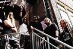 Valokuva Construcdeadin kokoonpanosta. Kuvassa näkyy lauma muusikkoja seisomassa teräskaiteen takana rappusilla. Osalla jäsenistä aurinkolasit silmillä ja kesäajan vaatetusta yllään.