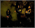 Synkkä valokuva Urn-yhtyeen kolmesta miespuolisesta muusikosta savuisessa luolassa. Miehillä pitkät hiukset, mustat vaatteet, piikkikoruja ja luotivöitä yllään.