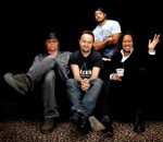 Metallica-bändin jäsenet istuvat mustaa taustaa vasten. Lattialla on jonkin villieläimen nahka miesten jalkojen ja tuolien alla. Kuvassa neljä bändin jäsentä, jotka hymyilevät.