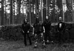 Ryhmäkuva Sadistik Forestin kokoonpanosta seisomassa metsässä muurin edustalla.