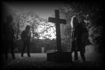 Mustavalkoinen valokuva Grotesque-yhtyeen kolmen jäsenen hahmosta, jotka seisovat hautausmaalla hautaristin lähellä. Taaempana on lehtipuita ja taivasta.
