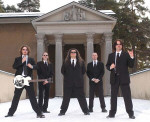 Candlemass-bändin miehet pukeutuneina tummiin pukuihin. Miehet seisovat lumisella tiellä. Taka-alalla komean rakennuksen oviaukko, jossa on neljä kivipylvästä.