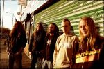 In Flames -yhtyeen jäsenet seisovat vihreästä aaltopellistä rakennettua rakennusta vastatusten. Kuvassa miehiä viisi kappaletta. Osa miehistä nojailee aaltopeltiseinään.