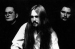 Mustavalkoinen valokuva Therion-bändin kolmesta jäsenestä, joista keskimmäisenä valkoiseen paitaan pukeutunut pitkähiuksinen mies, jolla viikset ja parta. Vasemmalla silmälasipäinen mies, oikealla mies, jolla viikset.