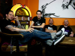 Volbeat-bändin neljä jäsentä oleskeluhuoneessa. Miehet istuvat. Osa istuu sohvalla, mutta vasemmassa laidassa näkyvä mies istuu tuolilla ja ojentelee jalkoja pöydän päällä. Hänellä on siniset farkut jalassa ja musta t-paita yllään. Käsivarressa tatuointej