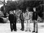 Mustavalkoinen promootiokuva Adastra-nimisestä bändistä, jonka jäseniä kuvassa näkyy viisi kappaletta. Miehet seisovat hietikolla, vasemmassa laidassa kaivinkone.