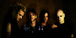 Valokuva Godhead-bändin kokoonpanosta, jonka jäsenet seisovat varjoissa. Kuvassa neljä miestä. Oikeassa laidassa bändin keulahahmo, jolla kalju valkoinen pää ja mustaksi meikatut silmänympärykset.