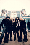 Nightwish-bändin promokuva vuodelta 2007. Kuvassa myös yhtyeen uusi laulajatar, Anette Olzon. Miehet ja nainen seisovat suurehkon rakennuksen edessä hiekkatiellä.