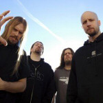 Meshuggah-bändin jäsenet seisovat sinistä taivasta vasten. Kuvakulma alhaalta ylöspäin, ruohonjuuritasosta ylöspäin, joten miehet näyttävät suurilta. Kuvassa neljä heppua, joista oikeassa laidassa näkyvällä ei ole hiuksia päässään.
