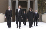 Candlemass-yhtyeen jäsenet saapastelevat lumisen maan päällä pukeutuneina mustiin pukuihin. Taustalla suuri rakennus, jossa valkoisia pylväitä oviaukon edustalla.