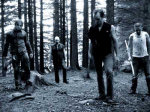 Sinisävyinen valokuva Manngard-bändin joukoista, jotka seisovat keskellä korpimetsää. Miehiä kuvassa neljä kappaletta. Oikeassa laidassa näkyvällä kourassaan kirve tai kuokka ja yllään valkoinen paita, joka on likainen.