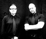 Mustavalkoinen bändikuva Deinonychus-yhtyeen kaksikosta, joista molemmat miehiä. Heillä yllään mustat kiiltävästä kankaasta valmistetut kauluspaidat, joiden kauluksiin solmittu samanlaisesta kankaasta ommellut solmiot. Miehet seisovat rivissä mustahkoa ta