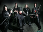 Viisihenkisen Domination Black -bändin mustiin pukeutuneet muusikot seisovat tummaa taustaa vasten. Kahdella miehellä kitara kourassa, yhdellä basso. Etummaisena seisoo silinteripäinen mies, jolla punainen leukaparta.