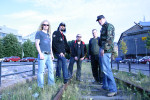 Sinisävyinen ryhmäkuva The Black League -bändin viidestä jäsenestä, jotka seisovat keskellä kaupungin autotietä sijaitsevaa viherkaistaletta, jossa kulkee junakiskot. Miehillä on normaalit arkivaatteet yllään, parilla lippikset päässä, osalla aurinkolasit