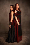 Kaksi Battleloren naista seisoo selät toisiaan vasten tummanruskeaa taustaa vasten. He ovat pukeutuneet pitkiin juhlamekkoihin, jotka ovat viininpunaisia ja mustia väreiltään. Molemmilla naisilla pitkät hiukset.