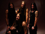 Neljä mustiin pukeutunutta Raise Hell -bändin miestä seisoo tummaa taustaa vasten ryhmäpotretissa. Miehillä samanlaiset asusteet kullakin: mustat hihattomat paidat ja mustat housut, jokaisella pitkät hiukset.
