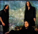 Kolme mustiin pukeutunutta Novembre-bändin miestä sinisävyistä koristeellista seinää tai verhoa vasten. Yksi etummaisena oikeassa alakulmassa sohvalla lojuva mies on parrakas. Muut kaksi miestä seisovat. Heistä jokaisella pitkät mustat hiukset.